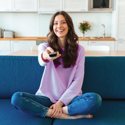 Image of a positive smiling optimistic young woman sit indoors at home watch tv holding remote control on sofa.