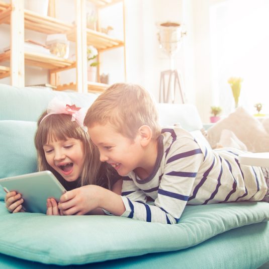 Happy siblings lying on sofa at home and playing with pc tablet together